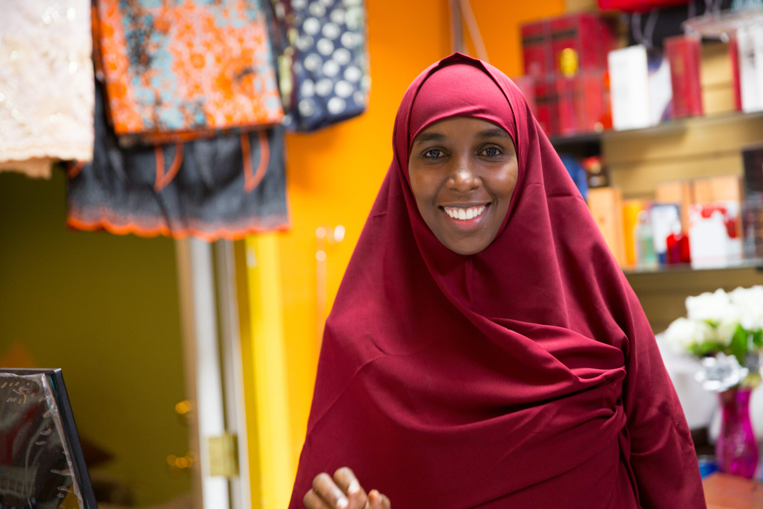 A smiling woman in a red headscarf stands in front of brightly colored store items.