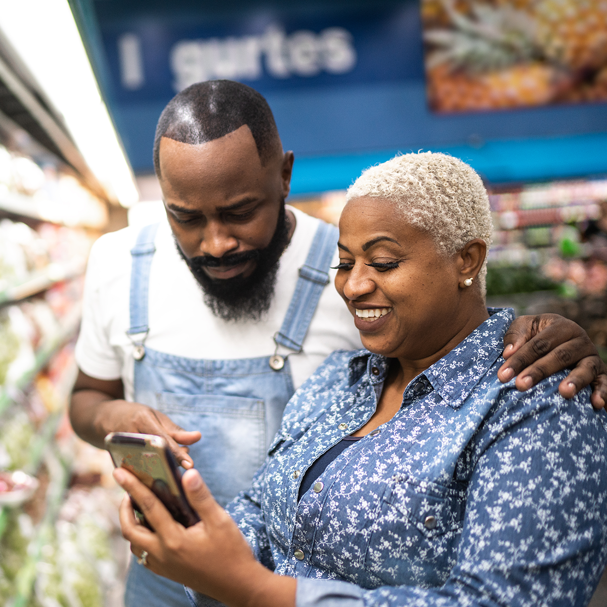 An African American man and woman in a grocery store smile while looking at a smartphone.